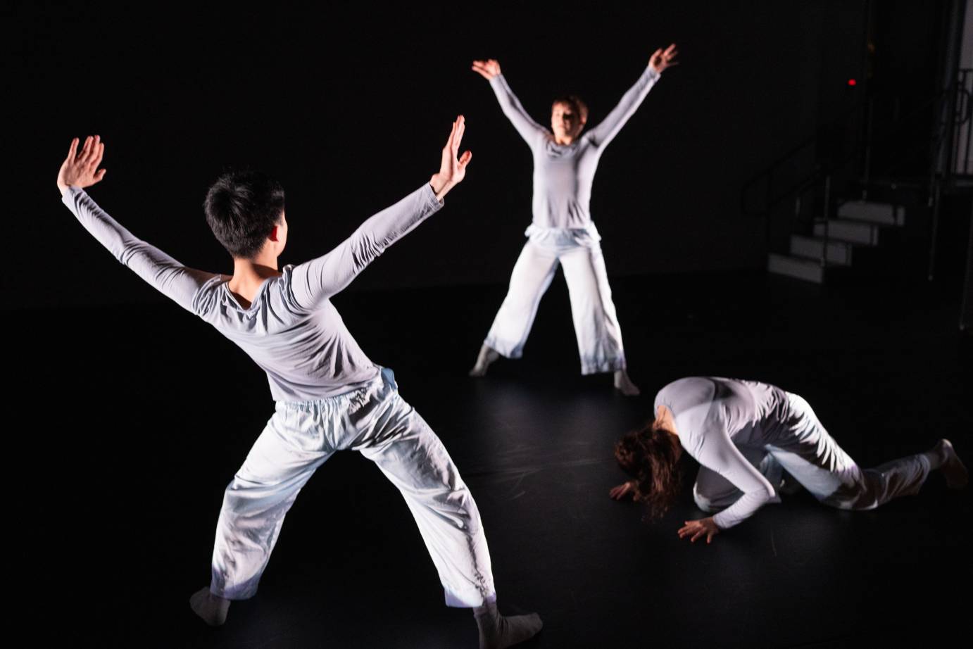 Three dancers in grey long-sleeved tops and pants, create a circle where two face one another in an X shape, while the third is on hands and knees, head tucked in, creating a contrasting ball shape.