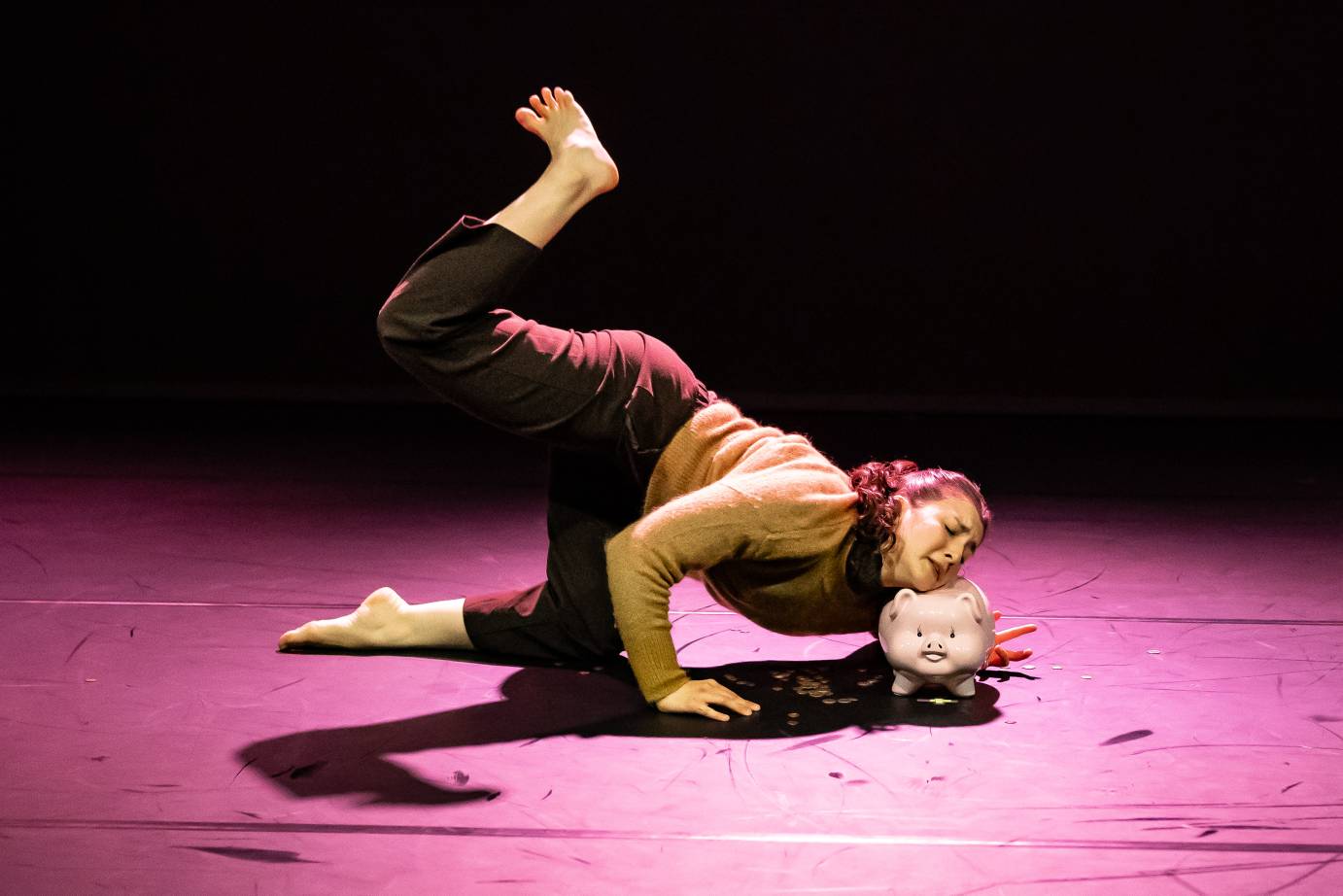 A woman, bathed in magenta light, kneels on one leg with the other reaching high, bent low to the ground and eyes closed, rests her chin on a piggy bank, 