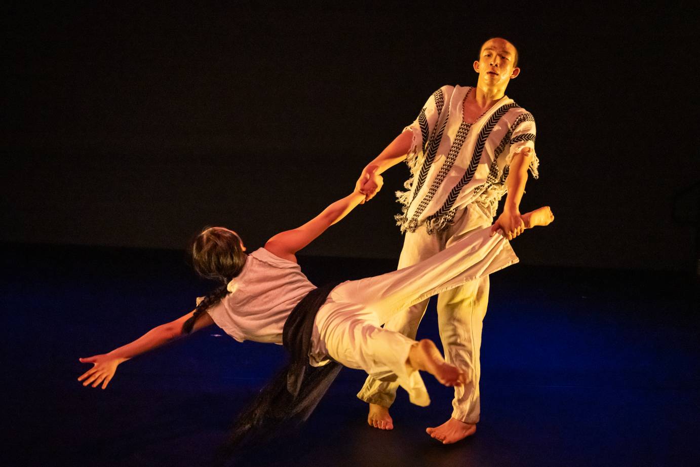A man in an Indigenous-looking top swings a woman in white top and black-belted pants by her arm and ankle.