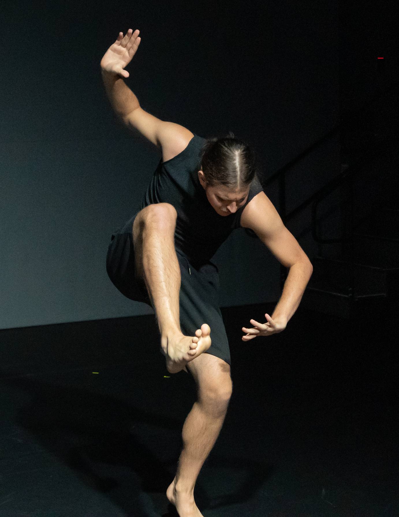 A dancer, wearing black shorts and tank top, with hair parted down the middle on bent leg, lifts the knee to their down-turned face, bent at the waist, arms reaching vertically. 