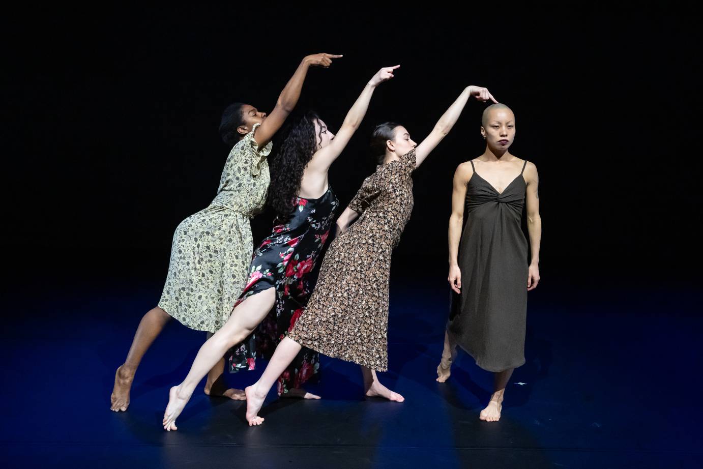 Three women dancers in flowered midcalf dresses point upward on the diagonal in profile, as they lean toward a bald-headed woman dressed in a brown spaghetti-strapped shift. The woman closest points her fingers atop the woman's head who faces foreword.
