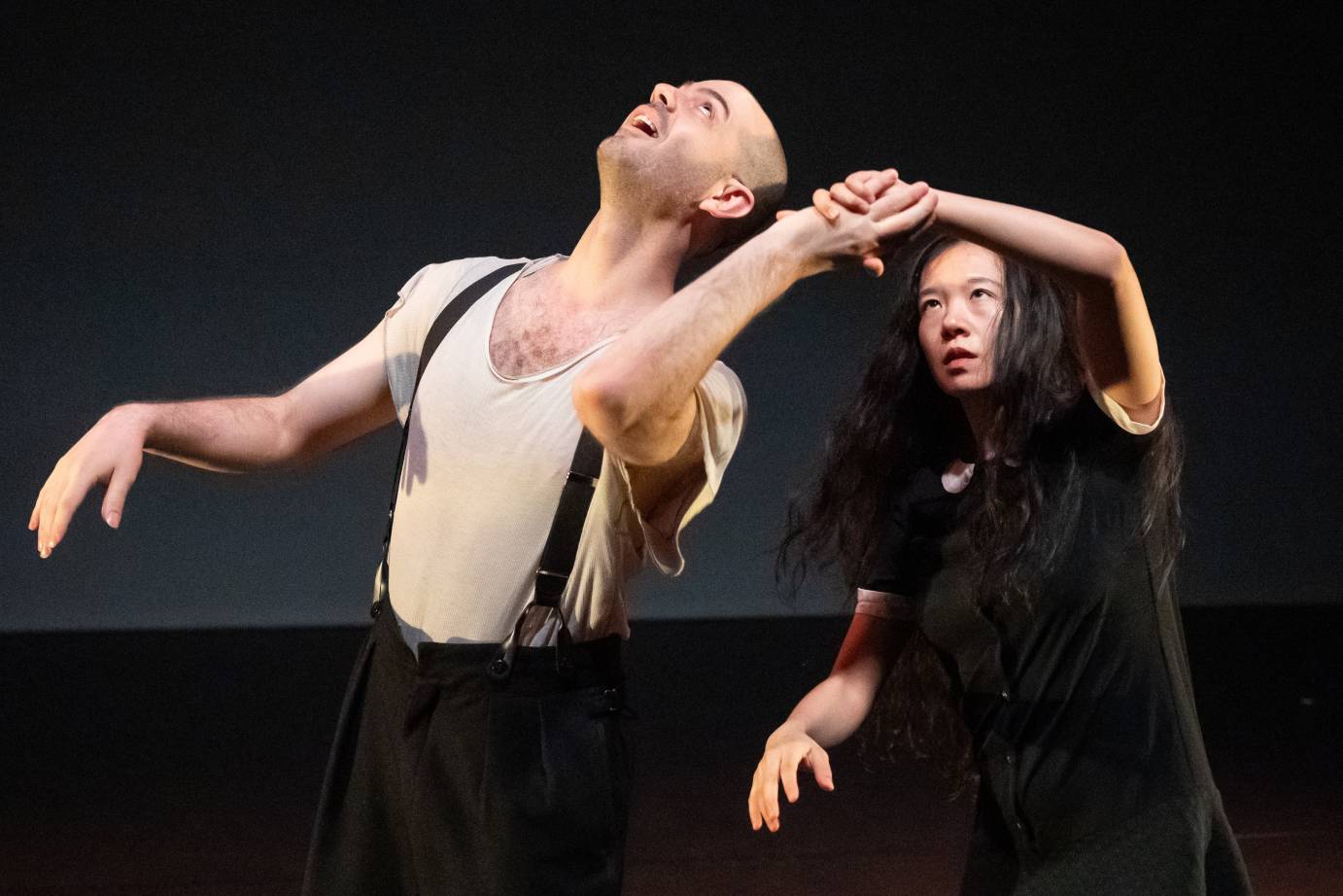 A man looking upward in a ragged white T-shirt and suspenders clasps one hand with a long-haired woman behind him dressed in black. He is smiling while her expression is serious.