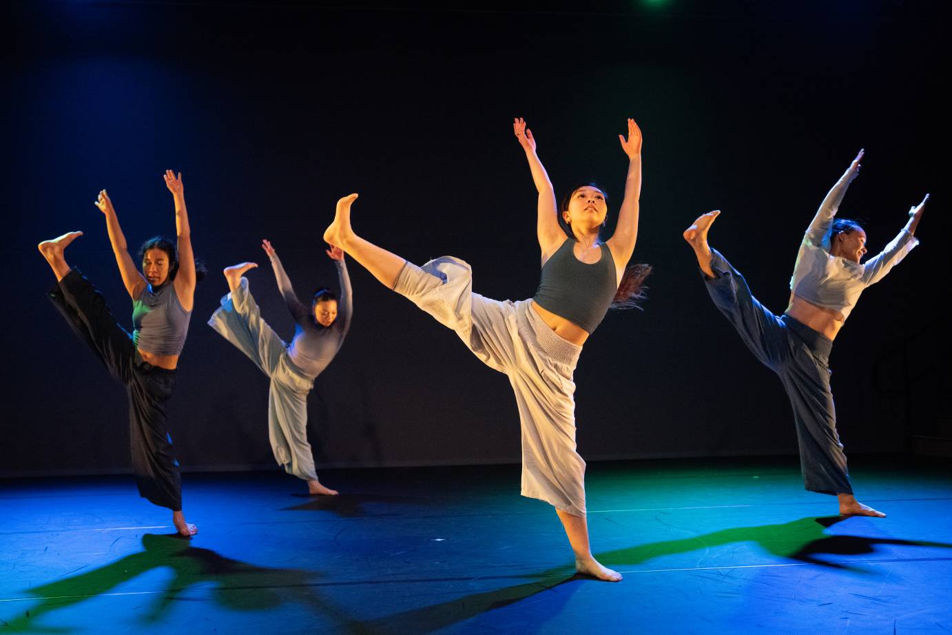 Four dancers in various tops and pants, facing front, reach their left leg on the diagonal and both arms high. Their heads are facing different directions.