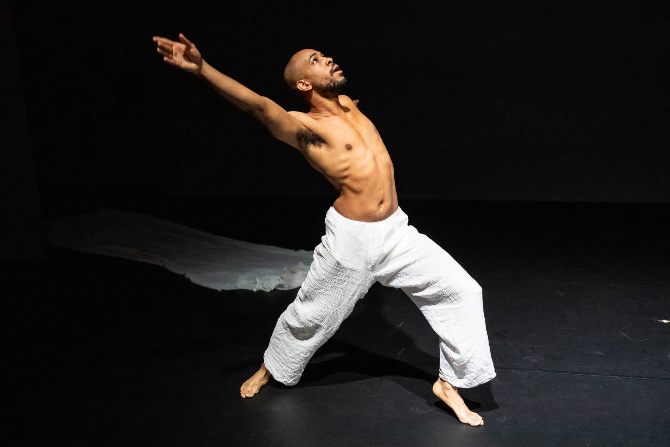 A bare-chested man in loose white pants opens his arms and body wide to the upper diagonal.