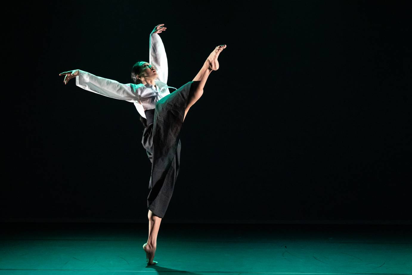 A woman dancer, wearing a loose white shirt and loose dark pants, on high releve lifts her right leg high to the ceiling head looking up and both arms tossed.