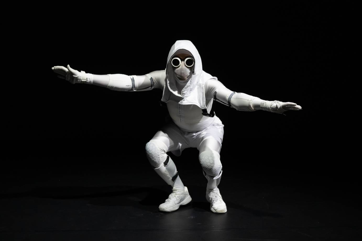 Figure wearing a head-to-toe white space suit and goggles stretches arms wide on the horizontal with feet planted and knees bent.