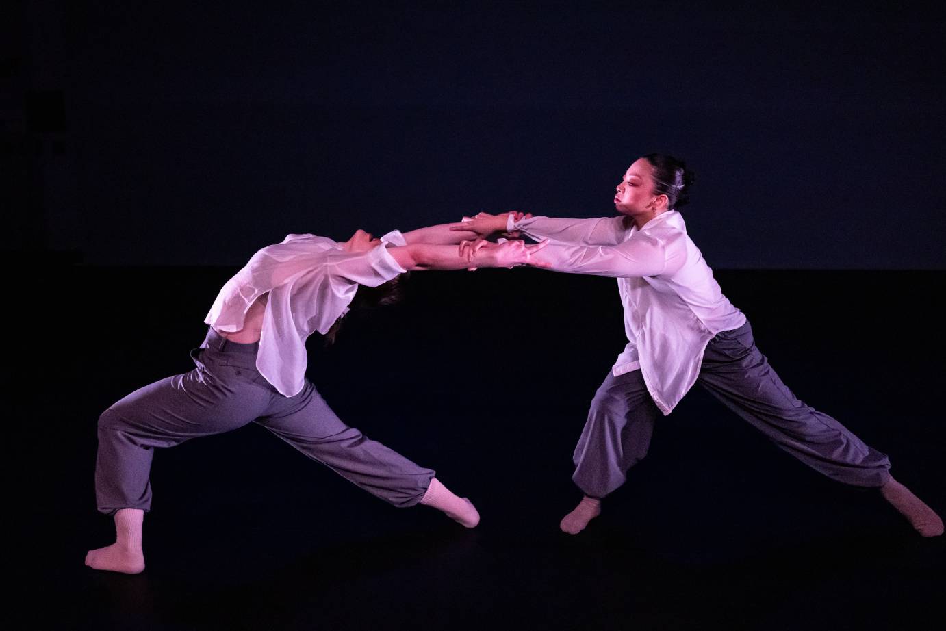 Two women dancers, dressed in gray slacks and white shirts, clasp forearms: one dancer in a lunge is bending backward while the other, also lunging, is facing her back.