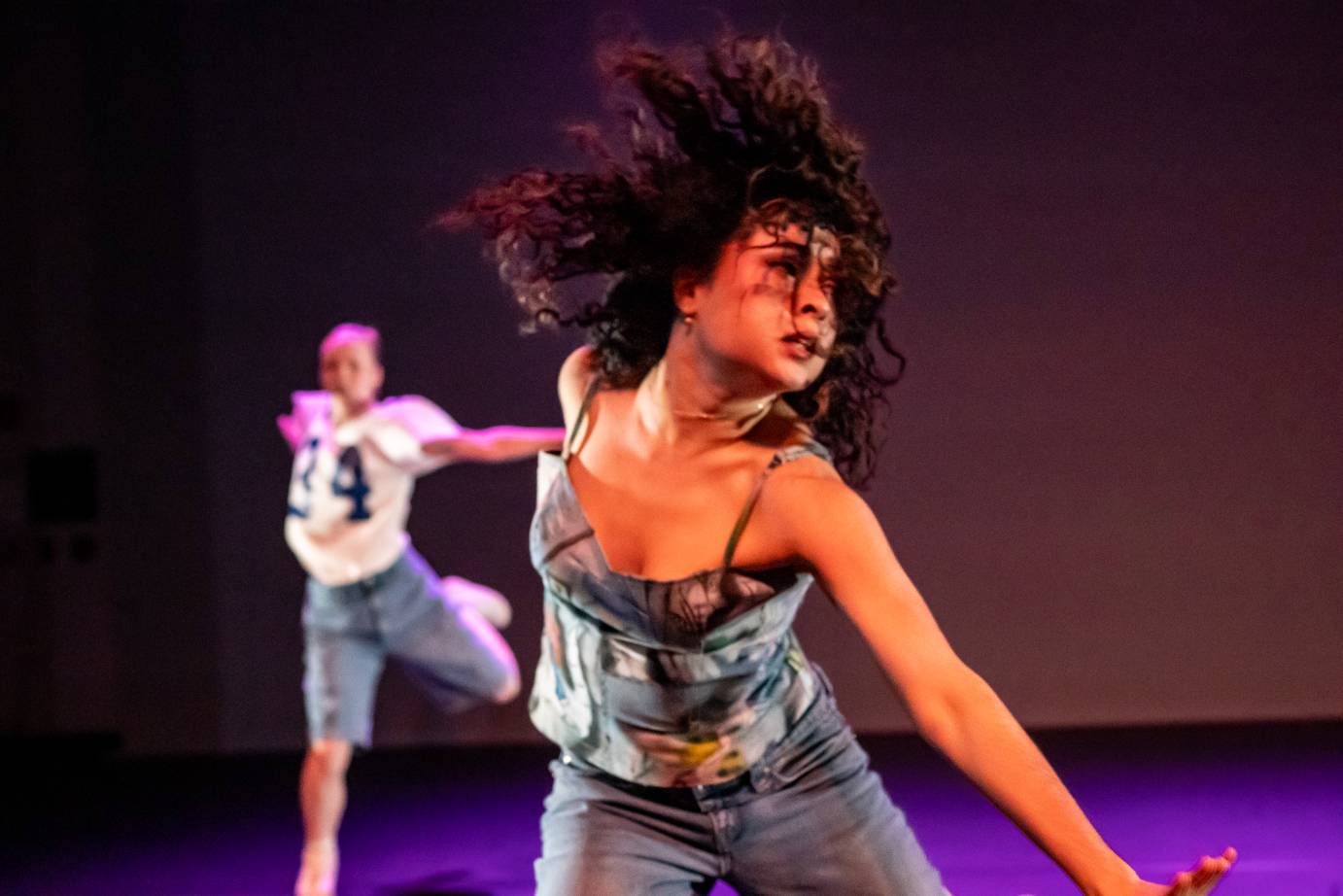 A woman dancer with shoulder length curly hair, strands of which fall onto her face, looks over her left shoulder as her body spirals to the left while her head looks over her left shoulder. She is pitched forward while standing wearing a spaghetti-strap top.