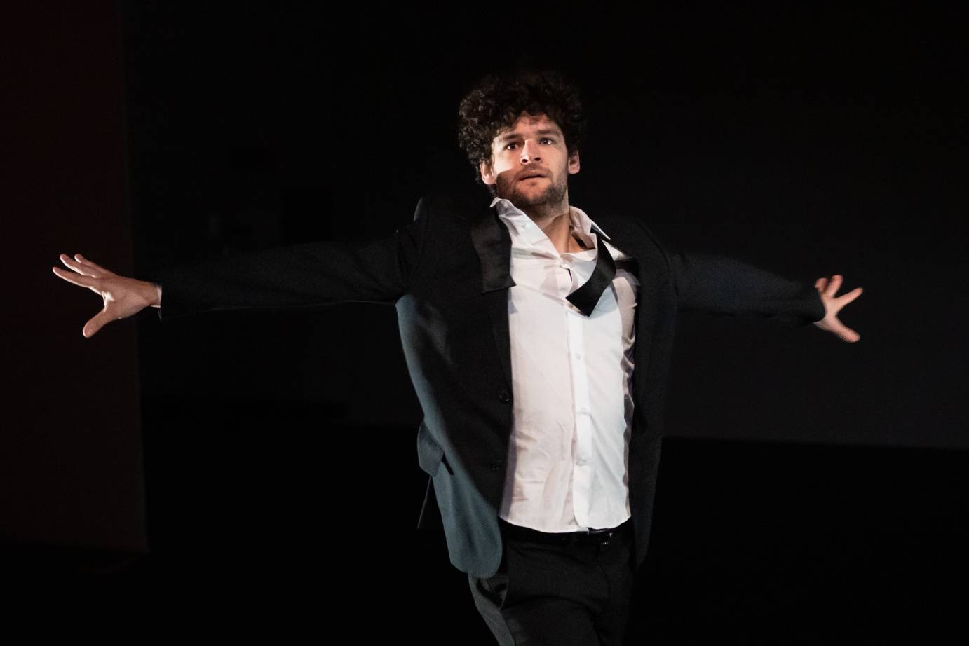 A man in black tuxedo with bowtie askew and white shirt open, stretches both arms horizontally, fingers splayed, looking over his right shoulder.