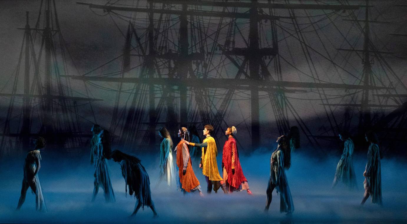 An illuminated woman in orange, a man in yellow and a woman in red, walk in serious-faced in profile. They are surrounded by ghost-like figures obscured by mist. 
