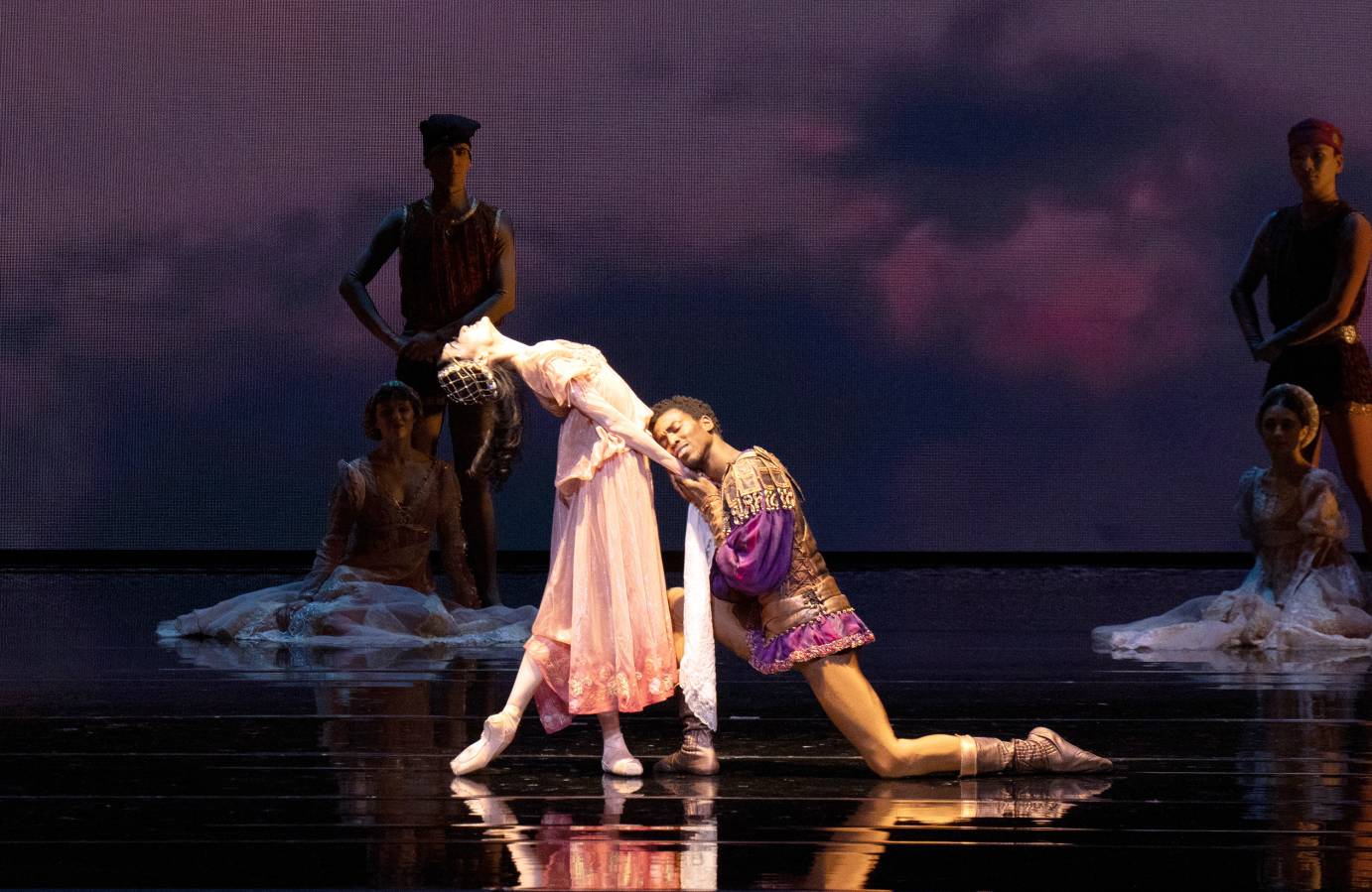 A standing woman costumed in a long mauve dress tilts her head backward, throad exposed to the ceiling. A man kneels at her feet, head resting on her belly, holding her downstage hand.