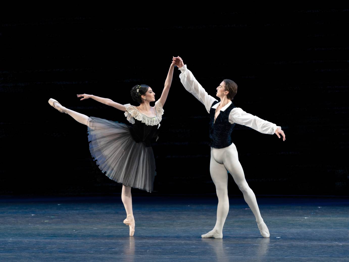 A ballerina in black dress and white flounces faces her consort dressed in white long sleeved shirt, black vest and white tights. They smile at one another hands held high above their heads. 