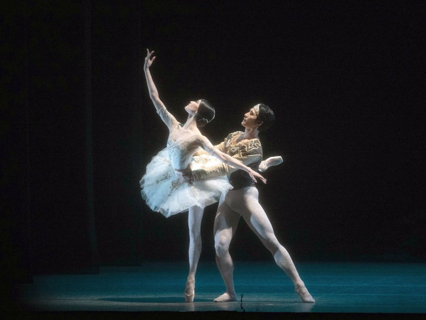 A dark haired ballerina costumed in a white tutu with arm and gaze upward on the diagonal, is held at the waist in back attitude by her partner dressed in white tights. His gaze follows hers.