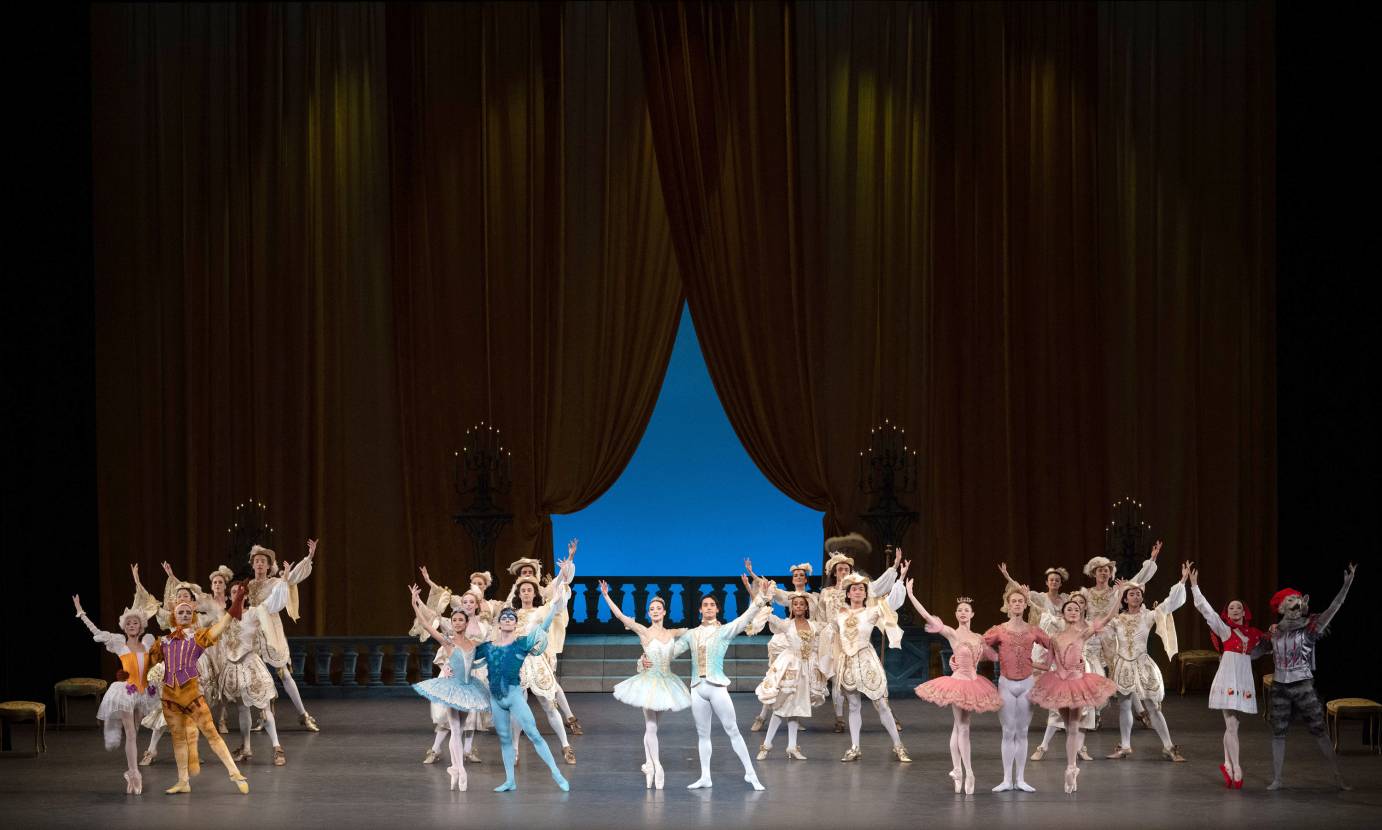 Against the rail of a balcony, with blue showing through an opening in dark curtains, the ensemble poses in roughly four lines. Two duets and a trio are slightly in front. They all raise their arms upward on the diagonal. 