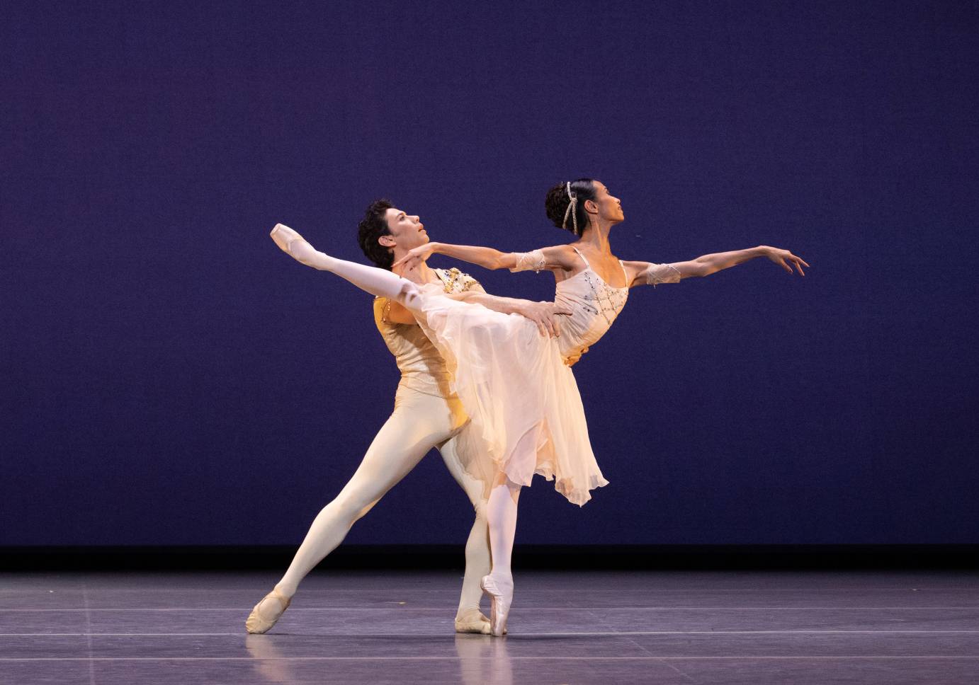 A ballerina on pointe and in arabesque with arms horizontal, wrists and fingers limp, is partnered by a man behind her hoding her at the waist.
