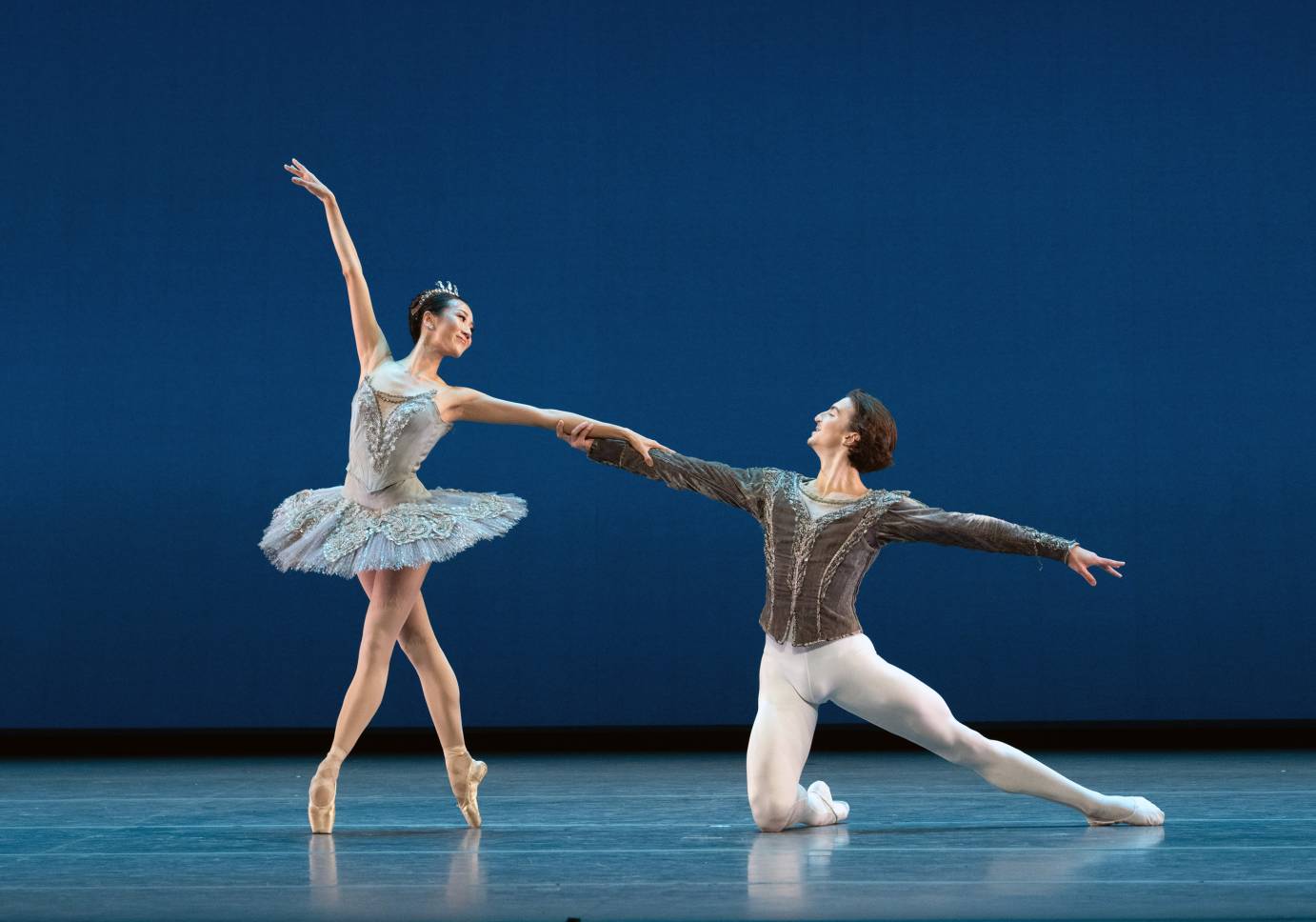 A ballerina, costumed in a light blue tutu, looks back and down at her partner, who is balanced on one bended knee with the other leg extended behind. They hold forearms while she is en pointe in an open fourth position. Her other lithe arm extended upward. 