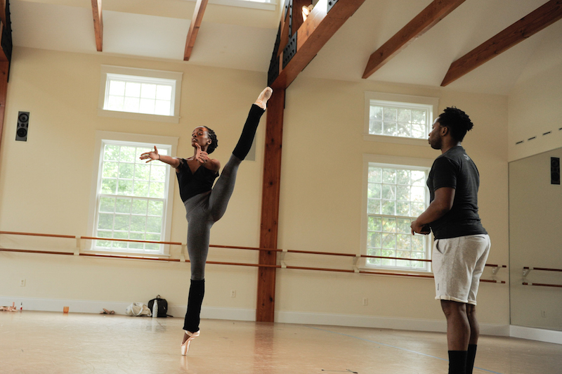 A woman lifts her leg high in the air. Her arms outstrech. Abdul coaches on the side.