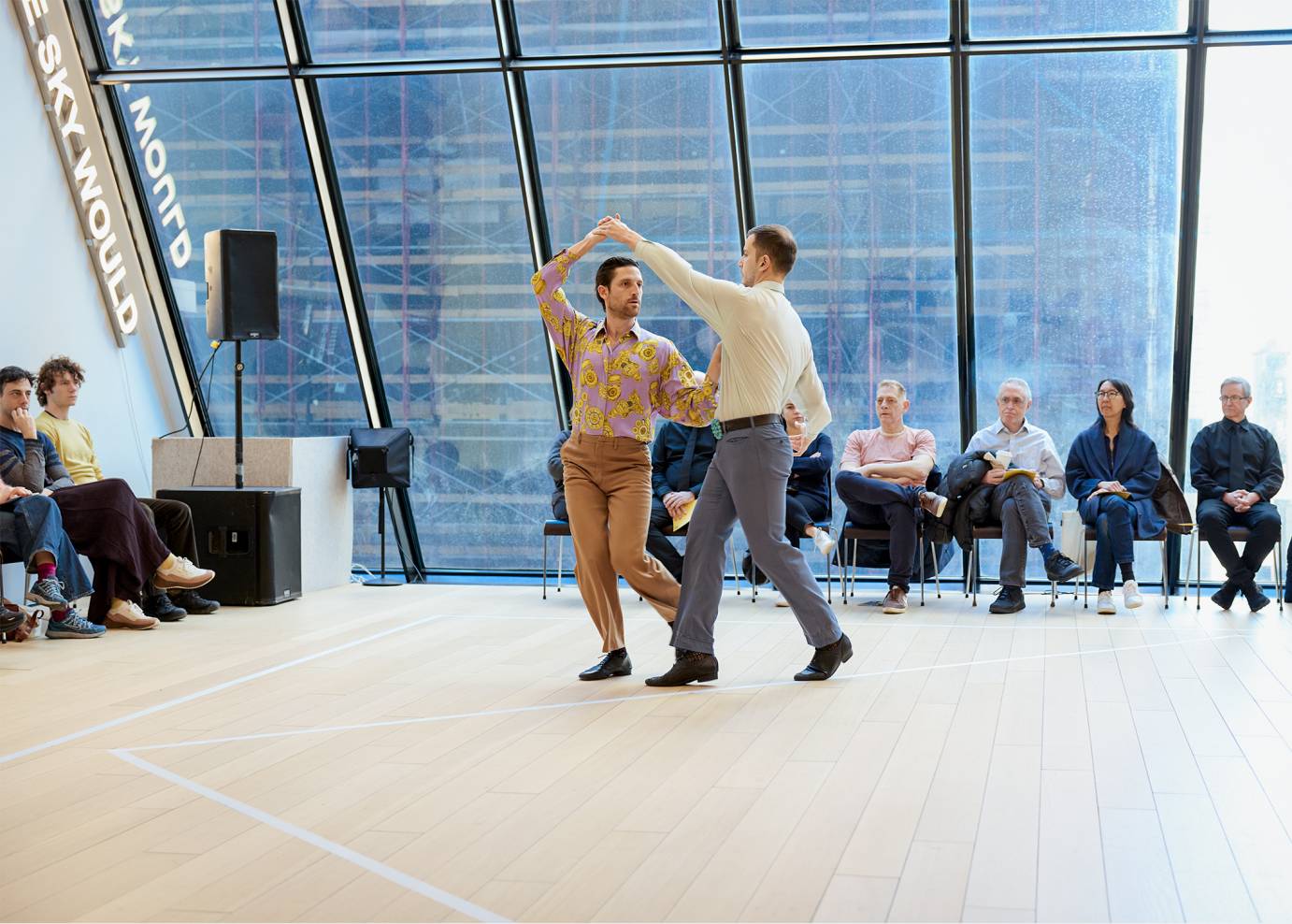Two men face one another while holding hands above their heads and below their hips against a glass backdrop with the city seen beyond.