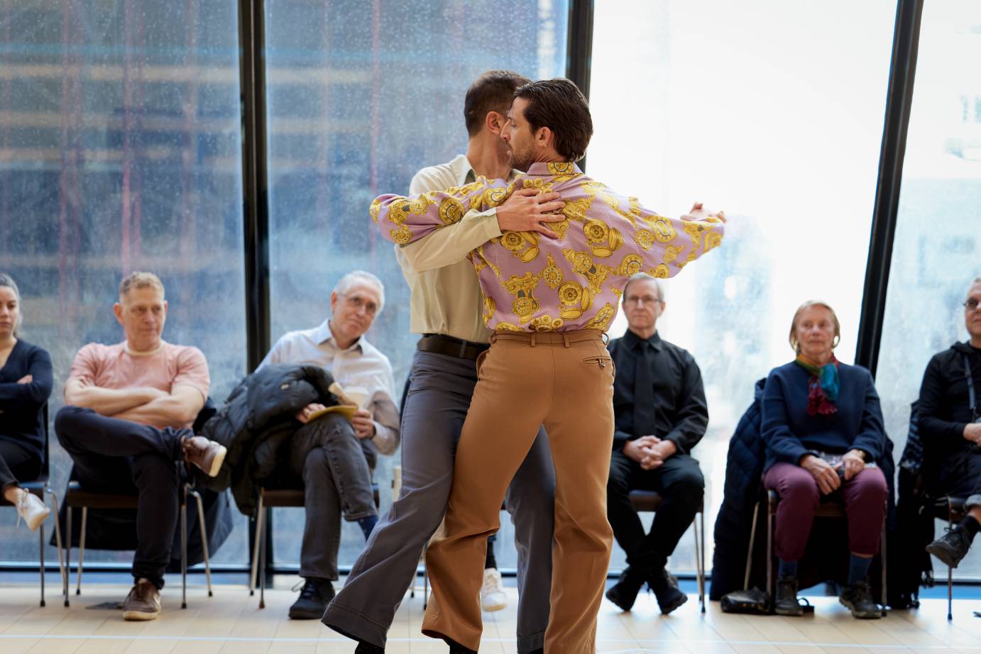 Two men embrace with heads turned in opposite directions. Arms enclose one another around their backs while their other arms are held loosely away from the torso, palms clasped.