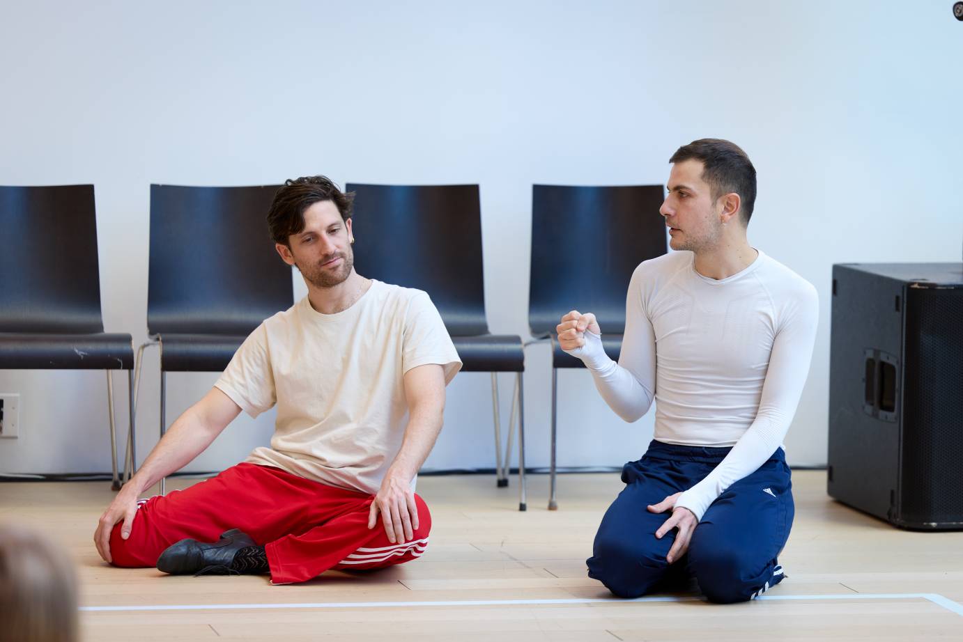 The dancers, in work clothes, sit side by side on the floor. One gestures while the other cocks his head and listens.