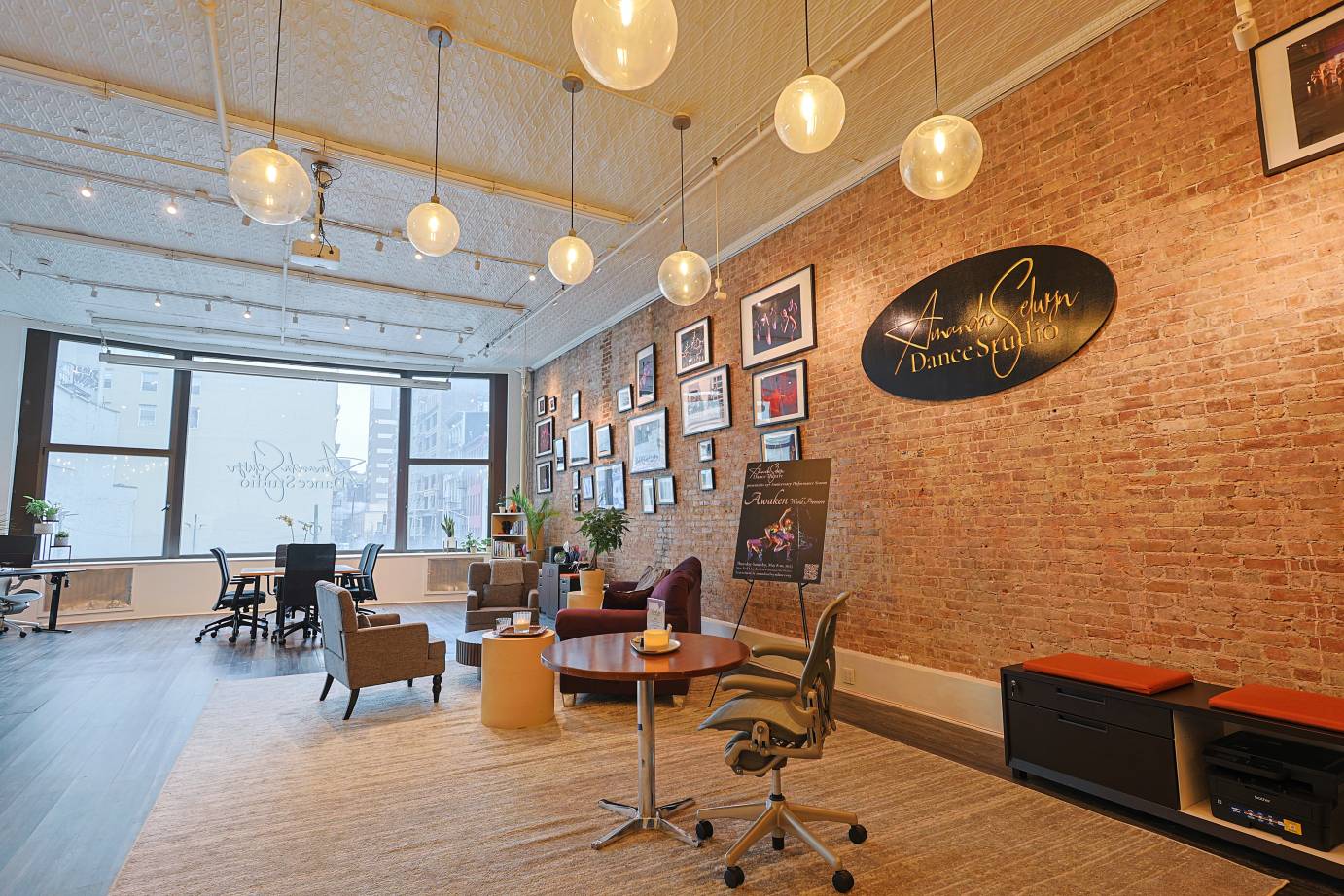 A view of a gathering place in the new studio with an Amanda Selwyn Dance Theatre plaque in black against a red brick wall and tables and chairs closer to large window looking out to a cityscape.