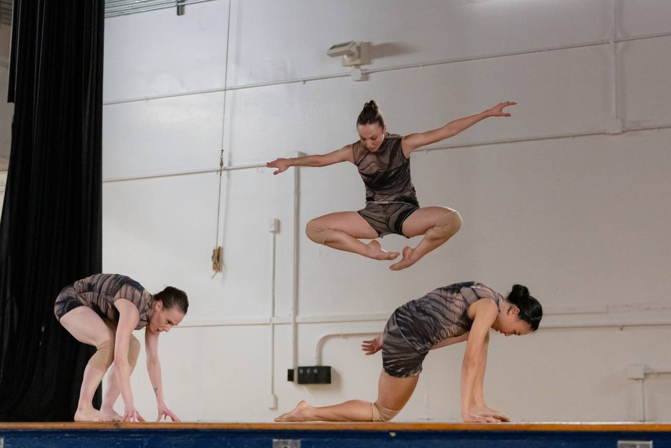 Three woman dancers in one piece gray and black swirled costumes: one dancer jumps off the floor turned out feet touching while two below are crouched.