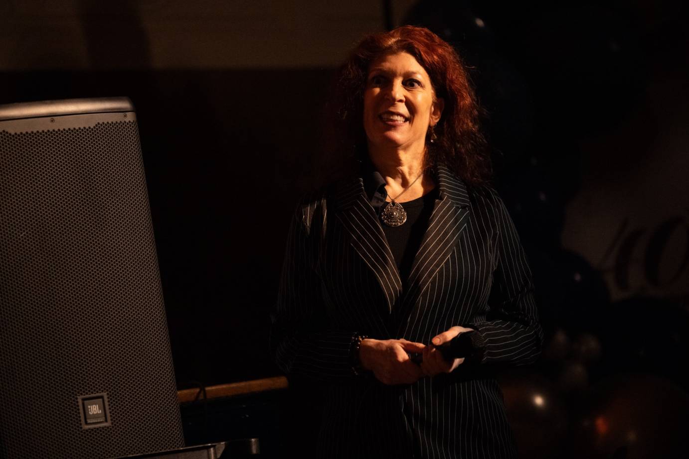 Amanda Selwyn - with shoulder length red hair wearing a striped suitcoat and medallion around her neck, smiles. 