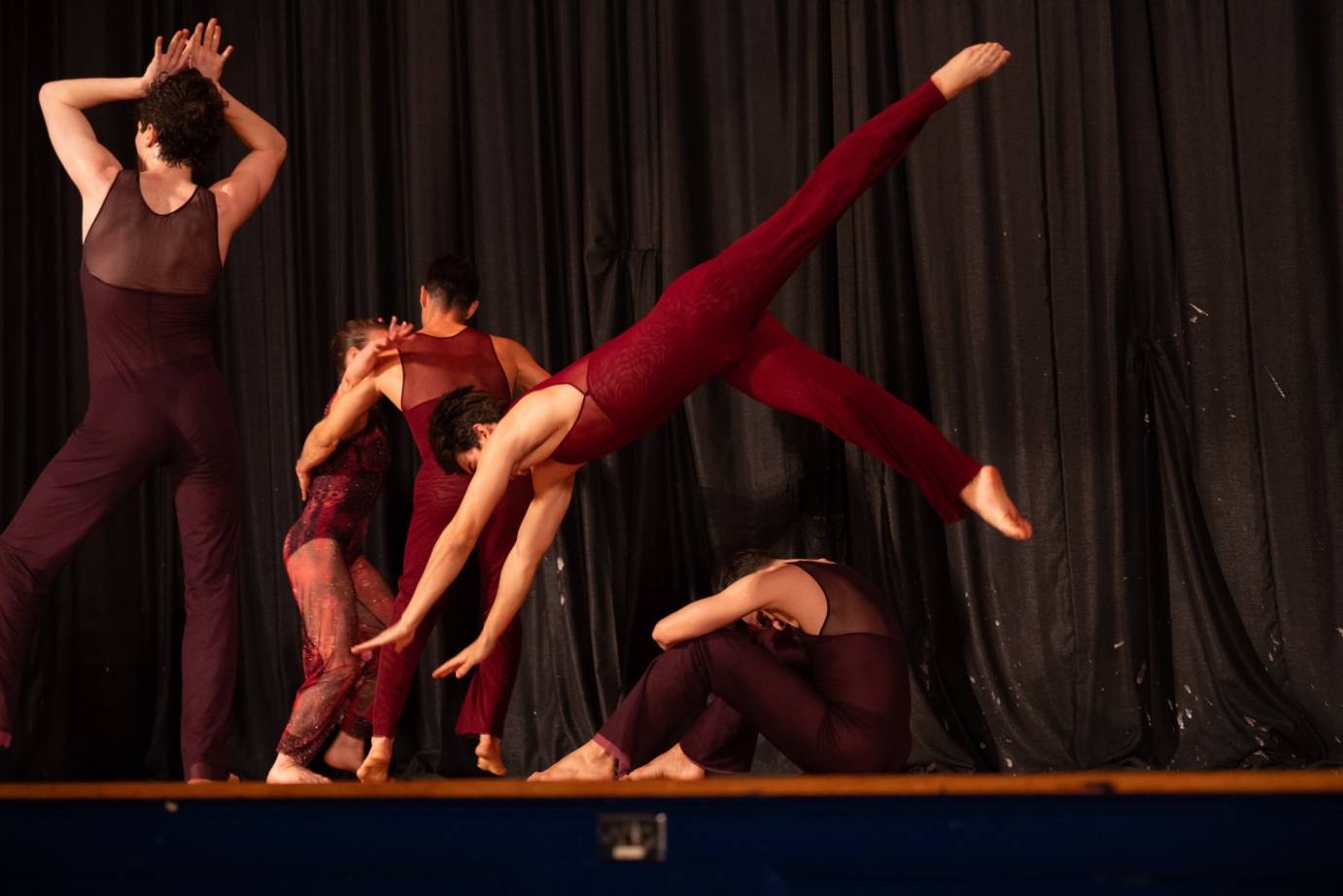 An airborne dancer in a red unitard-like costume dives to the floor - both arms are outstretched, one leg pointed to the ceiling the other handing below. Dancers are behind him sitting, standing and partnering, with their backs to the audience.