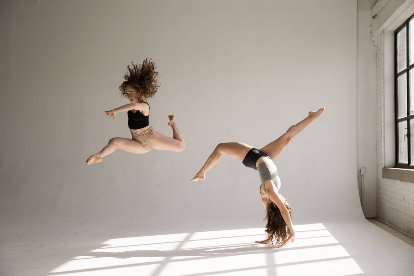 two women in midriff tops and beige briefs dance in a room against a white back drop... one is caught in mid-air her red curls flying above her, the other is in a handstand with her legs bent, you can feel their energy