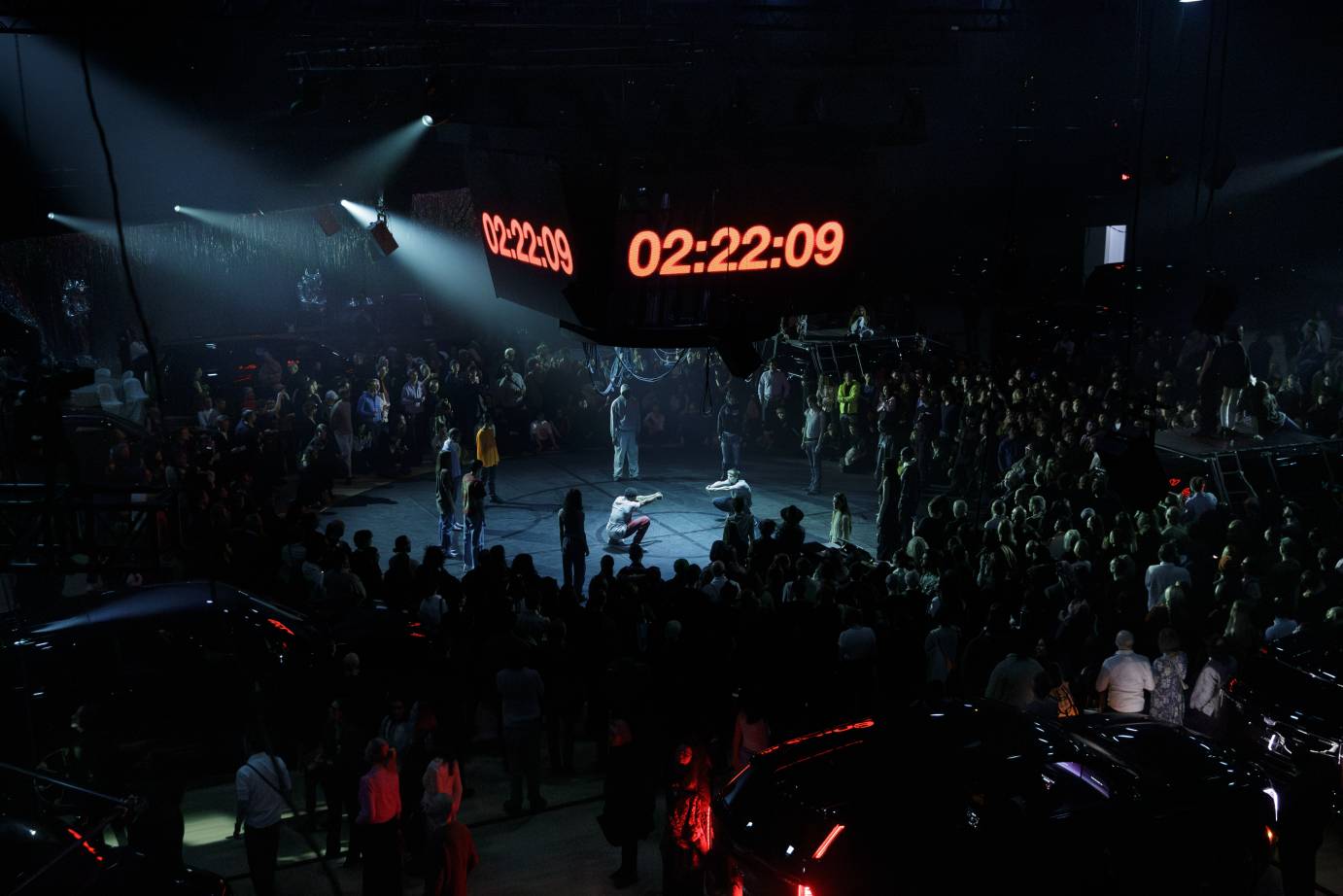 The jumbotron shows the time 02:22:29 in big red numbers under which a dance takes place. The audience, standing, surrounds.