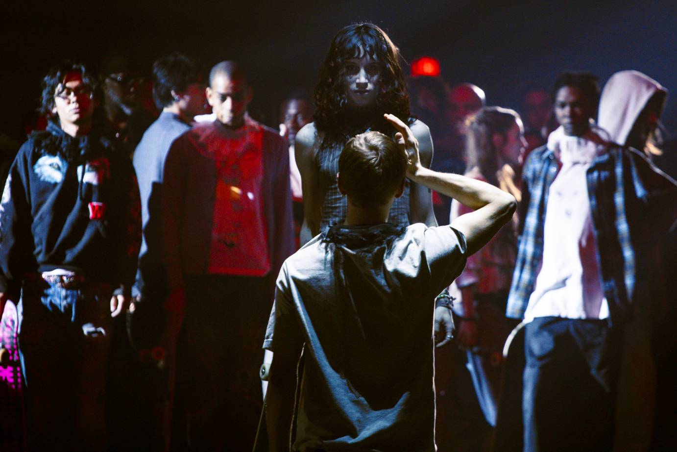 A crowd of youths in loose clothing including hoodies, sweatshirts, T-shirts stands facing a performer whose right hand is next to their head.
