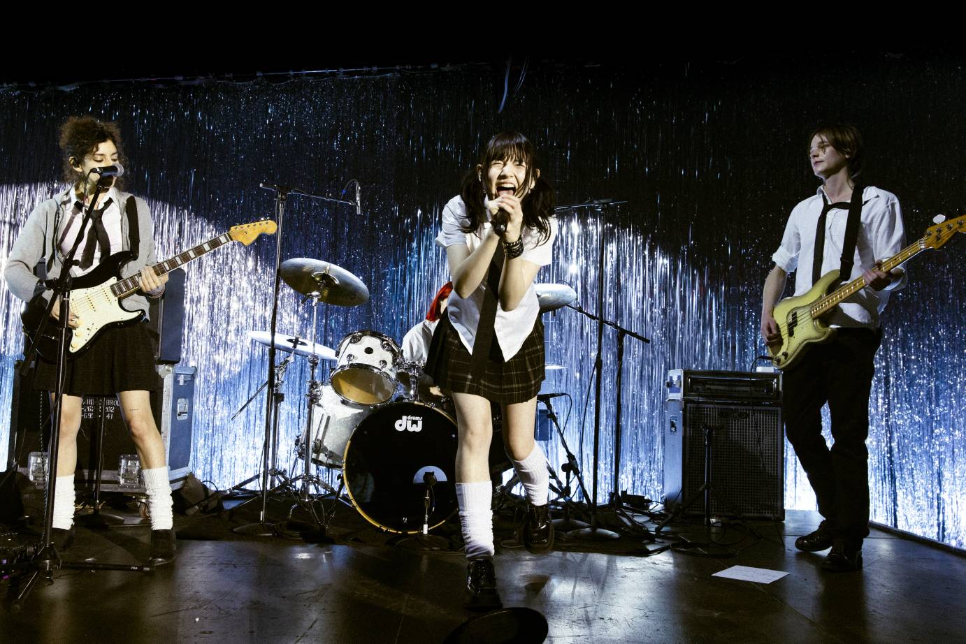 A four-person punk band, with a drummer and bass and guitar, surround the open-mouthed singer.