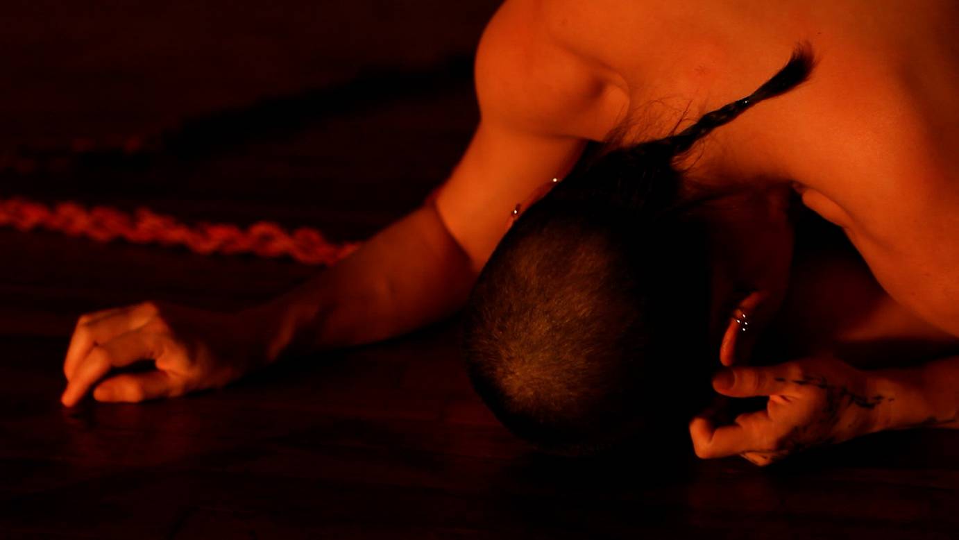 The back of an upper body - shoulders, arms and long, dark pigtail - bathed in orange light.