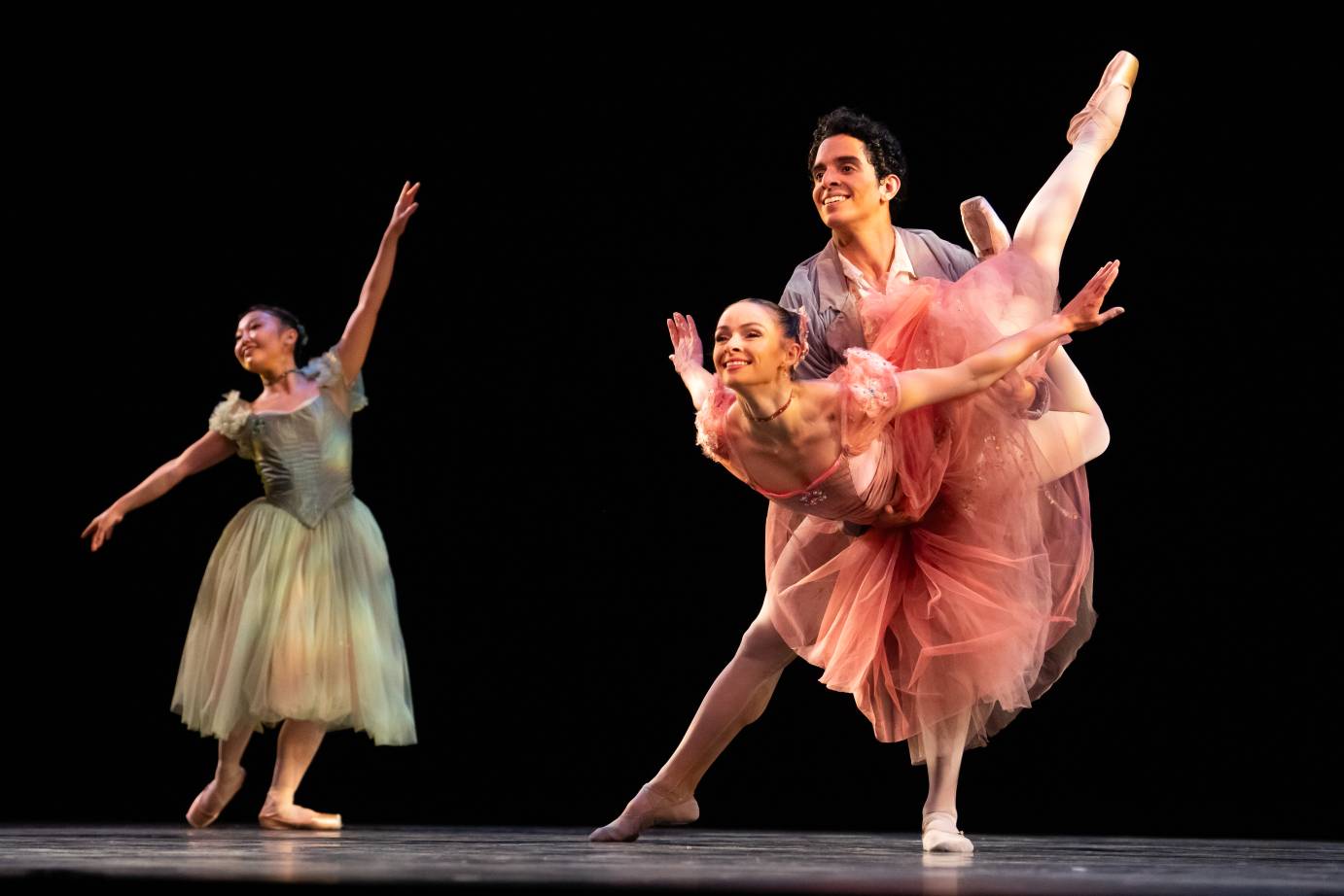 Three broadly smiling dancers face the diagonal: a ballerina is held at the waist by her swane, arms thrust back to meet her one long leg and one bent. A woman in bends her body slightly to the right with an uplifted left arm. 