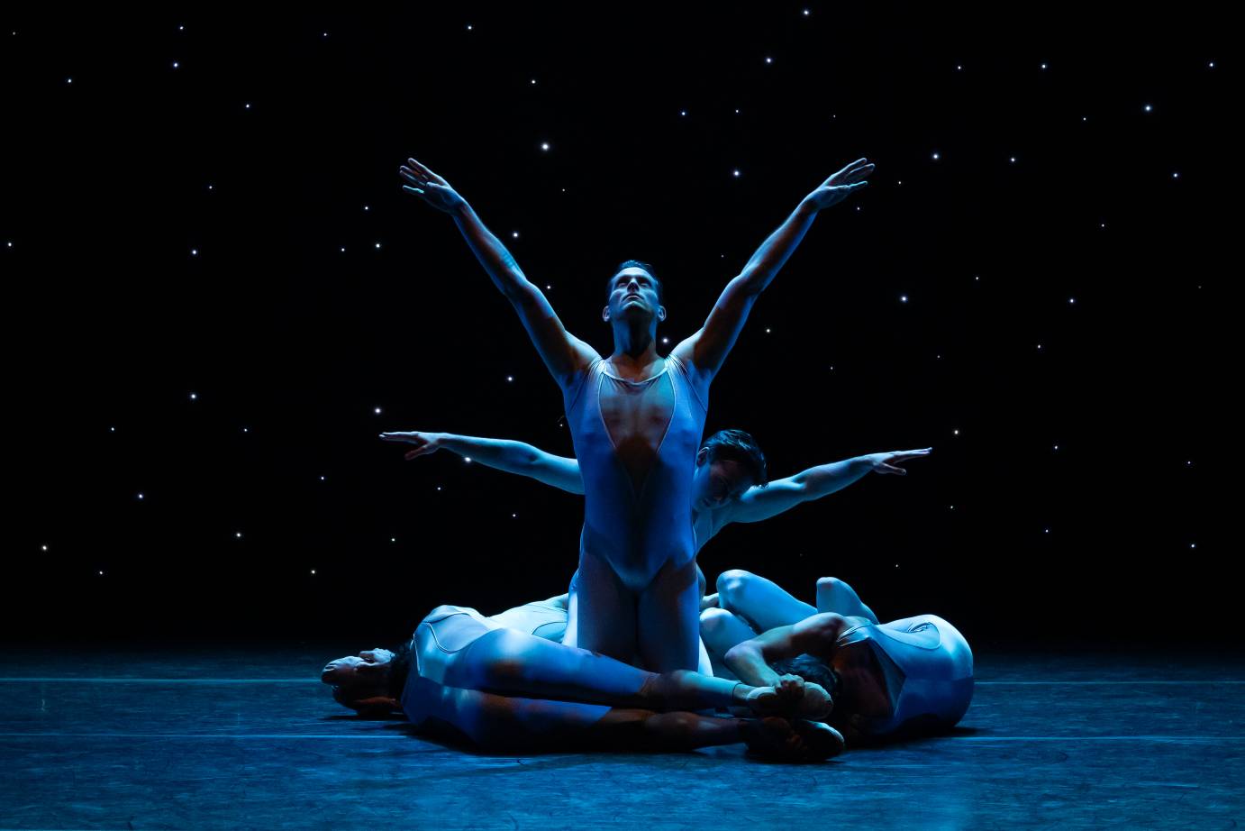 Bathed in blue lights in front of a background of stars, a man kneeling and facing forward with arms in a V above his upturned face, is wrapped by dancers at his feet and a dancer with winged arms behind.