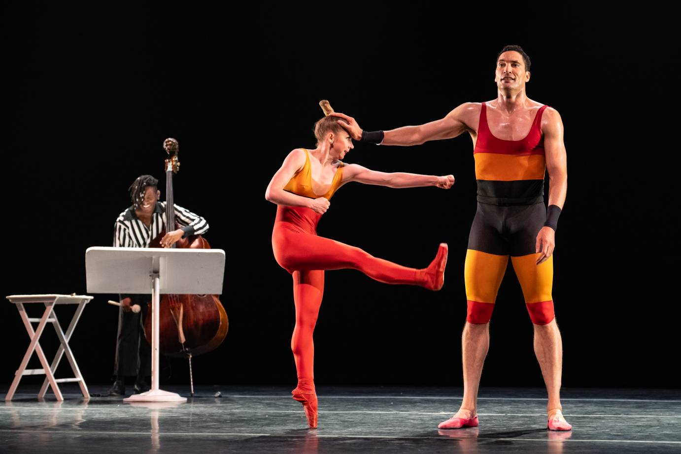 A bassist dressed in a black and white shirt plays behind a female and male duet. Dressed in red, orange and black the woman dancer tries to punch the very tall man who holds her away by placing his hand on her forehead. 