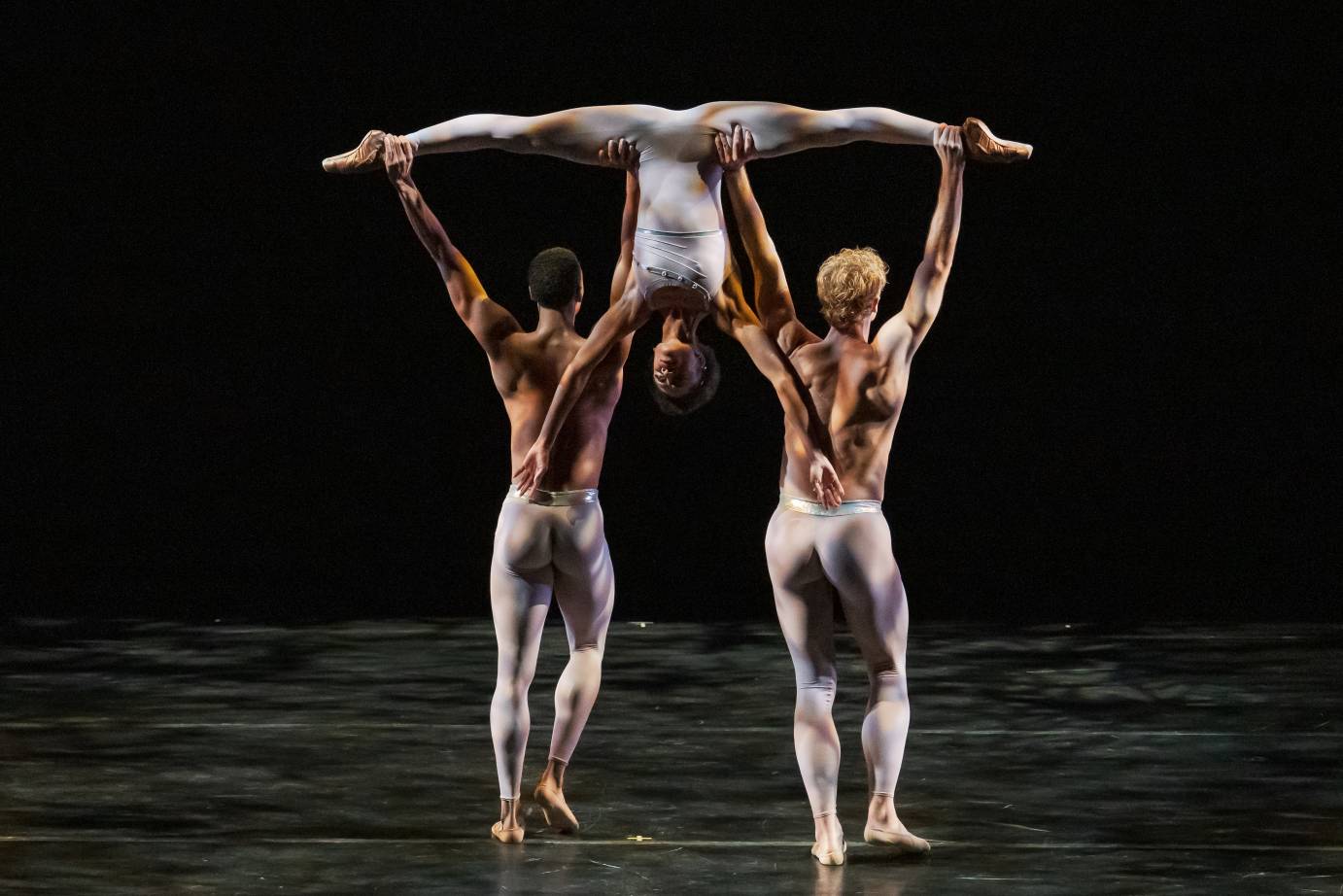Two male dancers hold a ballerina aloft upside down in a wide split. The men are bare-chested and wearing tights, their backs to the audience. The ballerina looks out her arms streaming down each man's back.