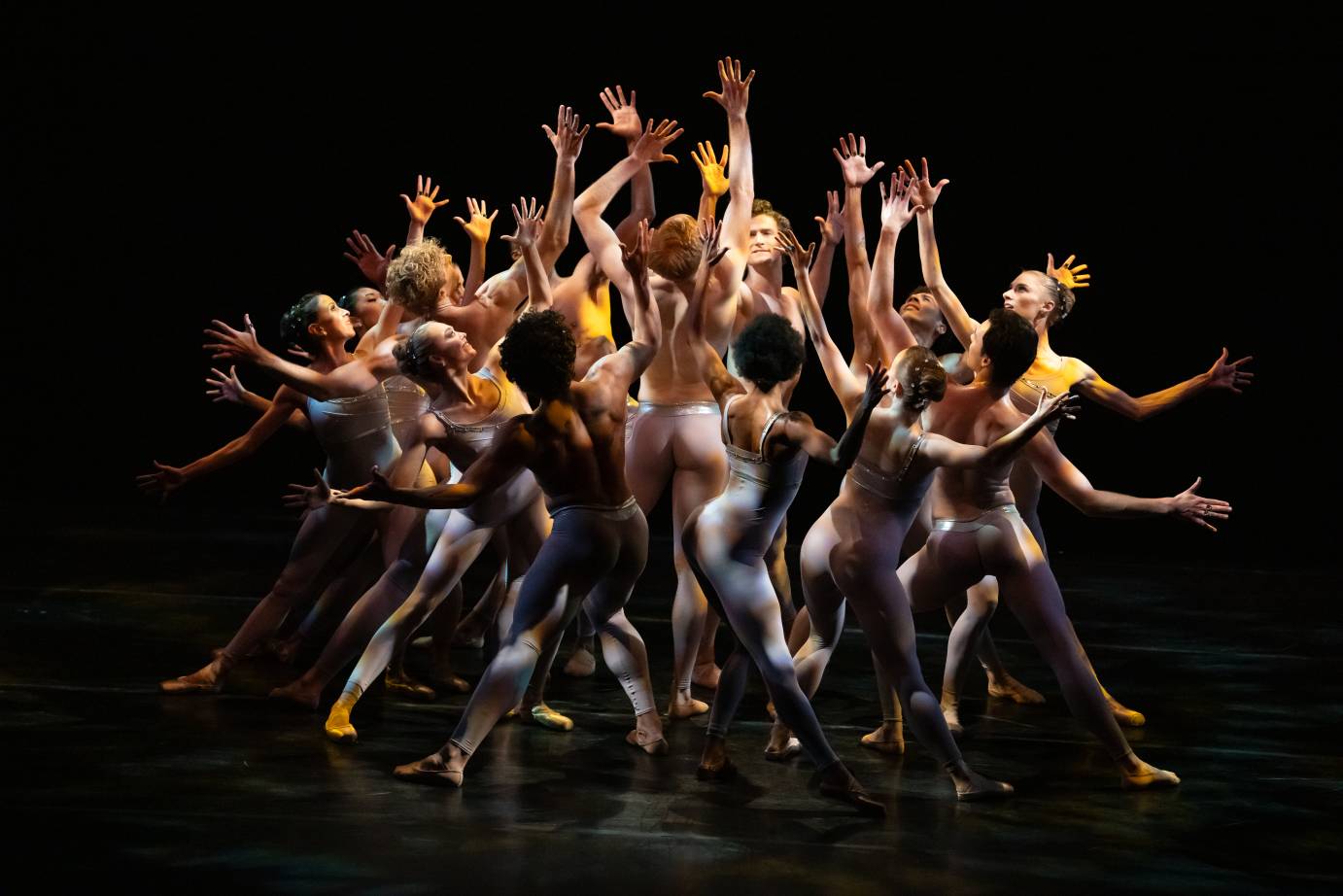 The ensemble of dancers cluster together, lunging and leaning away or standing tall, and reach their hands to a center point.