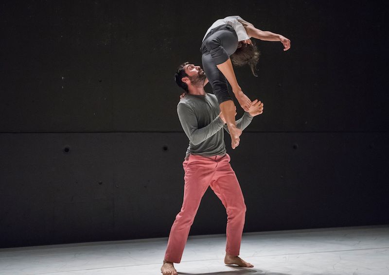 A woman does a back bend in mid air. She supports herself by pressing her hand into her partner's upper back. The partner spots her from below. 