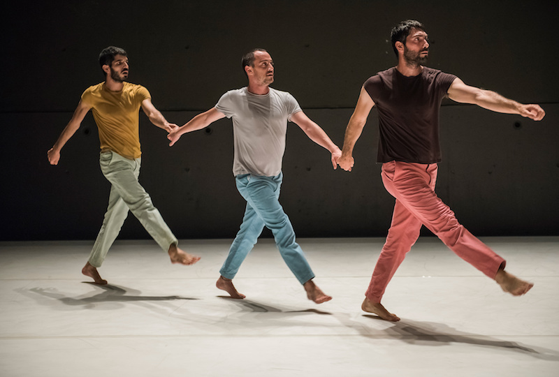 Three men in colorful t-shirts and chinos hold hands in a diagonal line. Their right leg is outstretched in front of them.