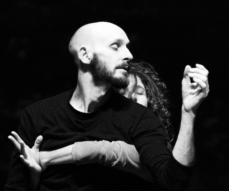 A woman with curly hair wraps her arms around Marc Brew. We see his face in side profile and his arms gesture out in front of him.