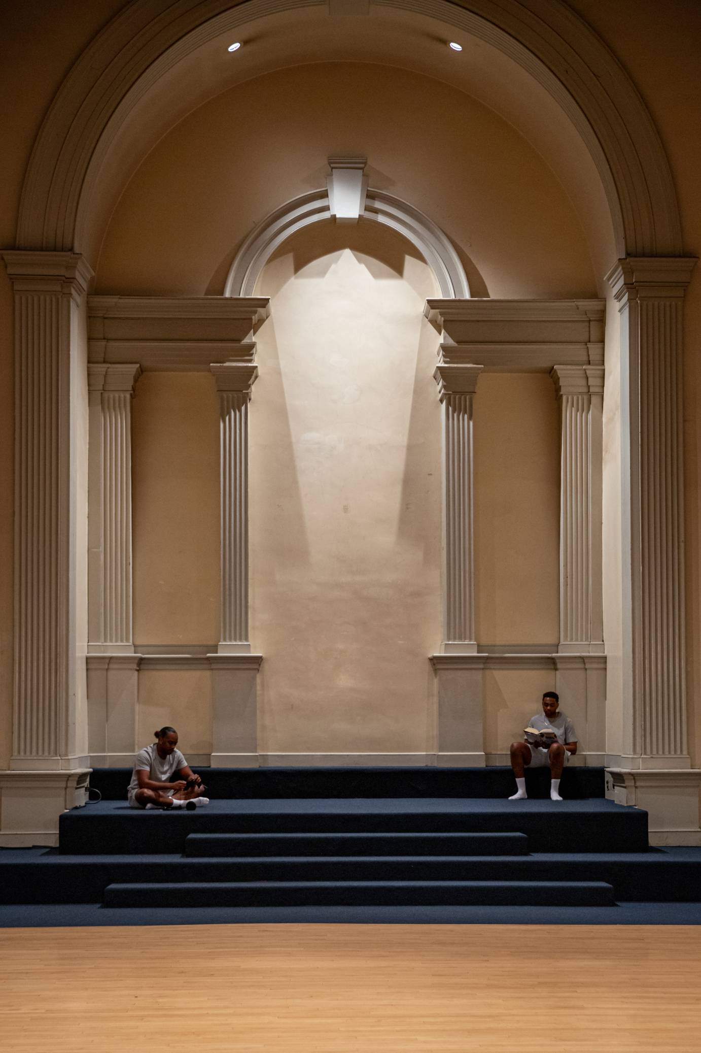 Two men sit on gray stairs in the alter of a church. One, cross legged, knits while the other perched a small ledge, reads a book.