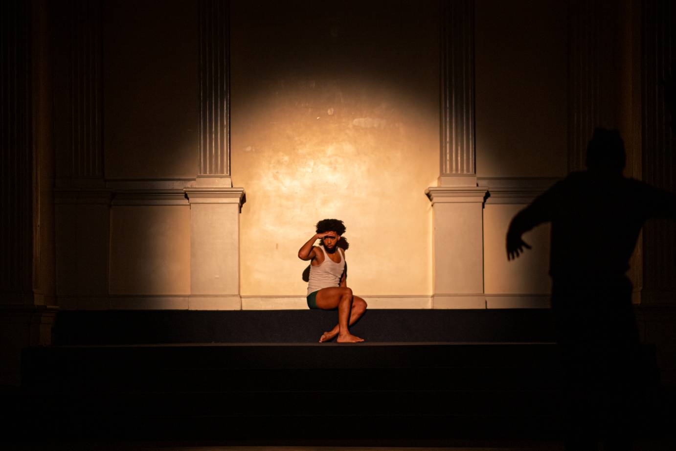 A man in shorts and T-shirt shielding his eyes sits in a spotlight.