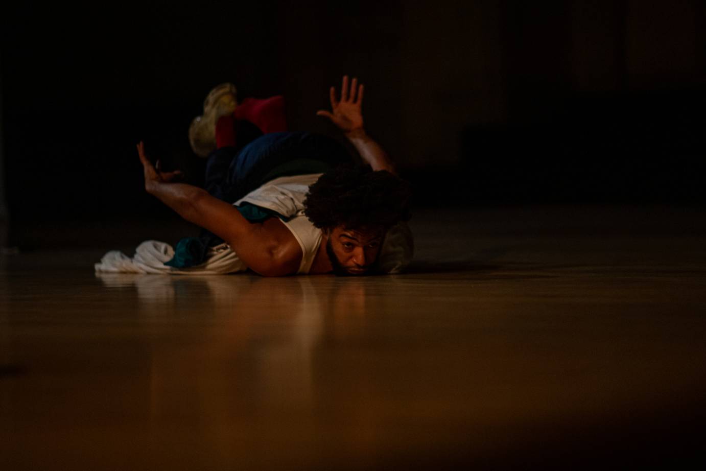 A man, dressed in a white sleeveless shirt, with the front of his body on the floor lifts his pelvis and the bottom of his feet skyward.