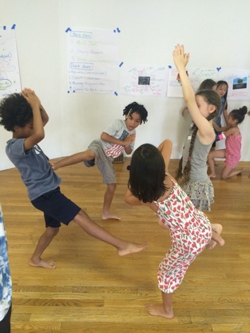 A group of kids play. One has his leg in the air. Two children have their palms together with their arms raised above their head. Another stands on one leg.