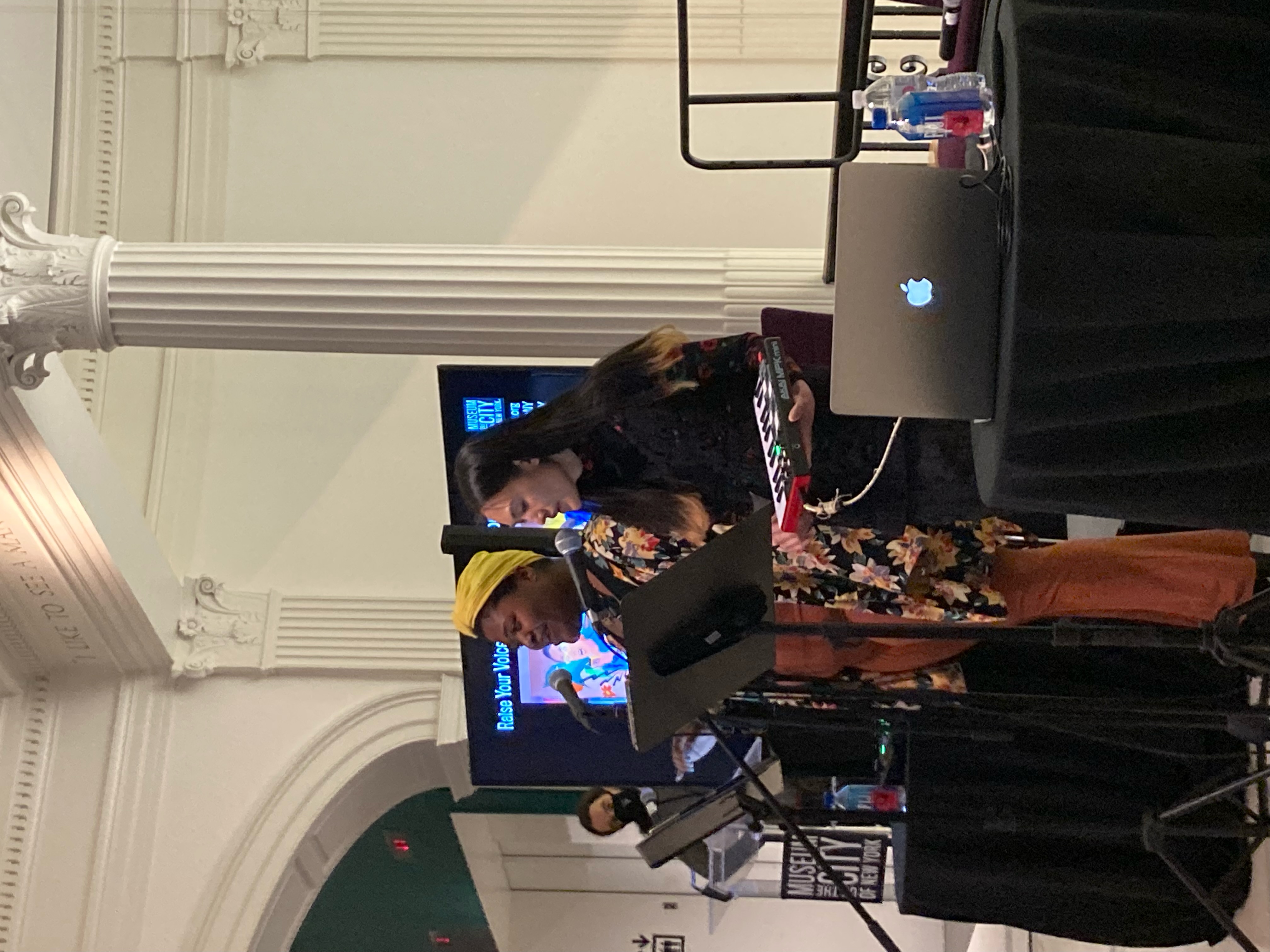 two women singing at mics, one Black with a yellow head wrap the other lighter skinned with long dark hair with blonde ends... she is holding a mini keyboard