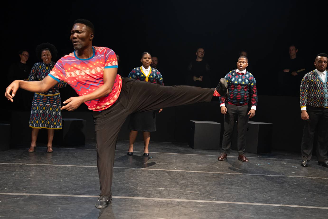Dancer in a bright red shirt stands in front of a group of singers, his left leg extended to the side