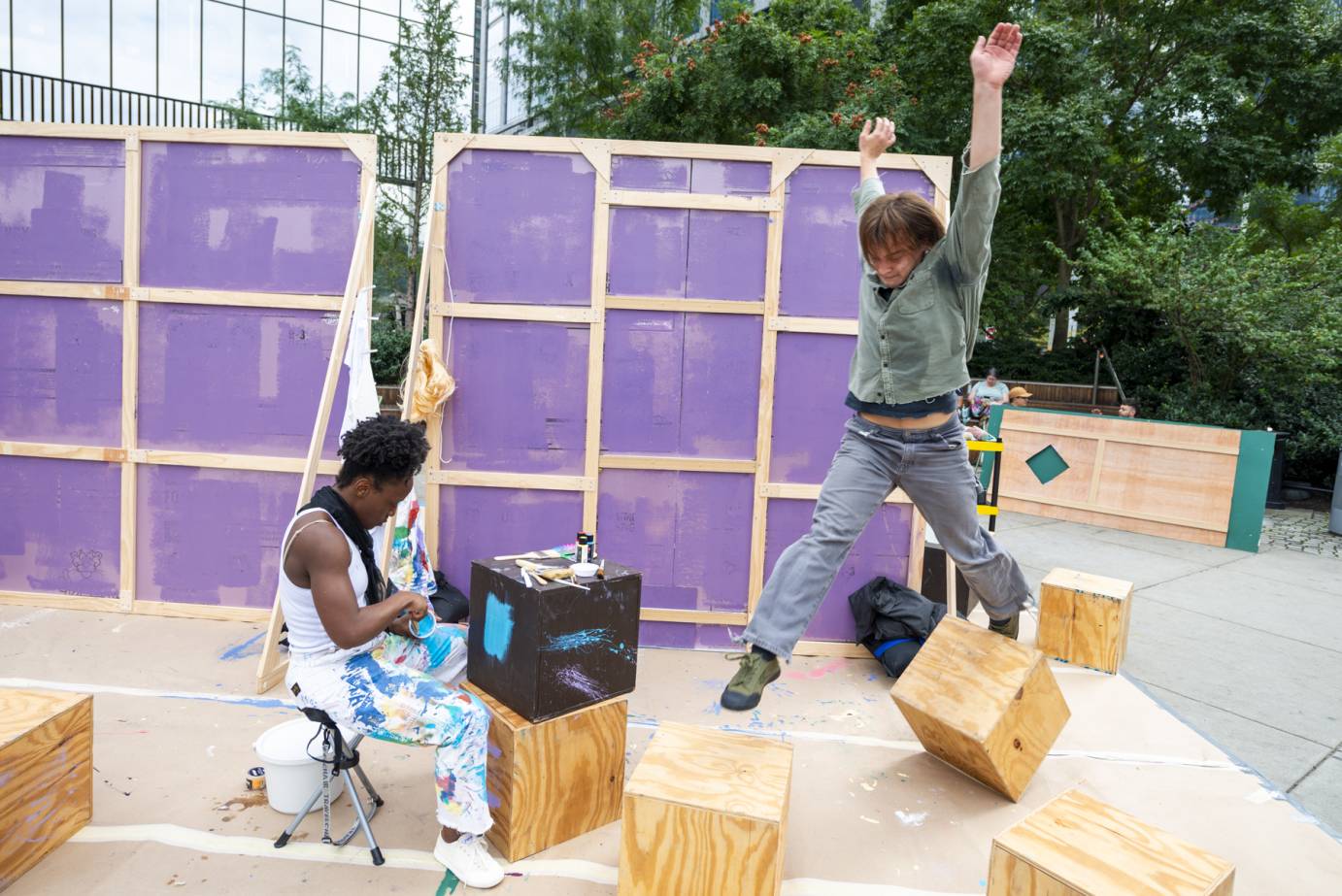 male dancer leaping over a cube. female sitting behind 2 stacked cubes mixing paint