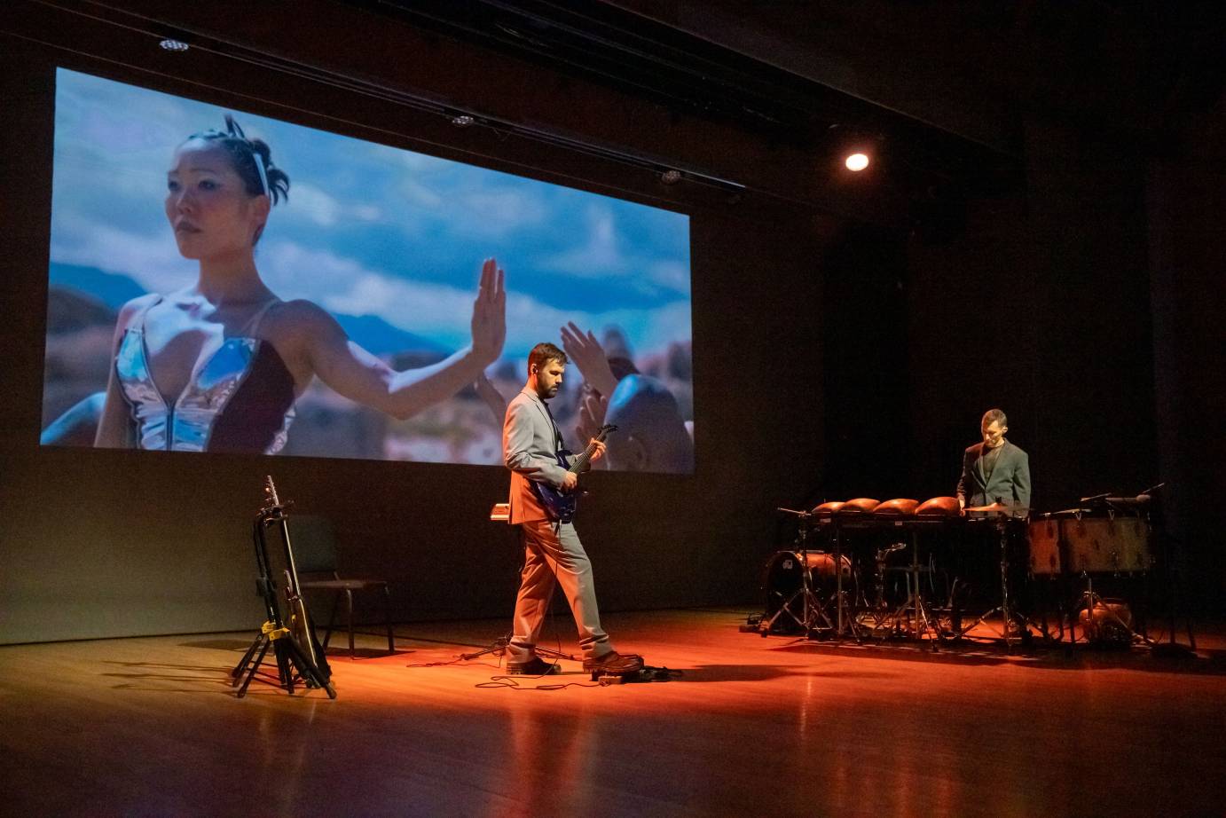 guitarist and drummer onstage in front of projection where dancer stands facing diagonally to camera, one arm outstretched to side and palm flexed