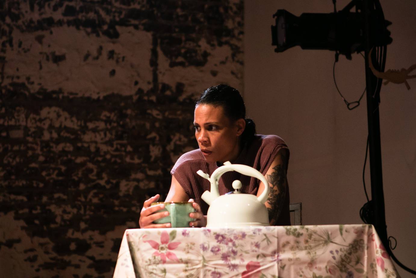 woman at a table holding a cup of tea in her hand. Tea kettle on the table in front of her. floral tablecloth